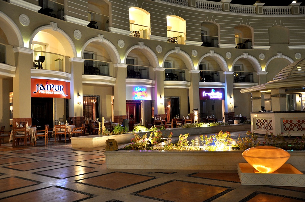 KaiSol Romance Resort Sahl Hasheesh (Adults Only): An illuminated resort courtyard with restaurant entrances, outdoor dining tables, and landscaped planters.