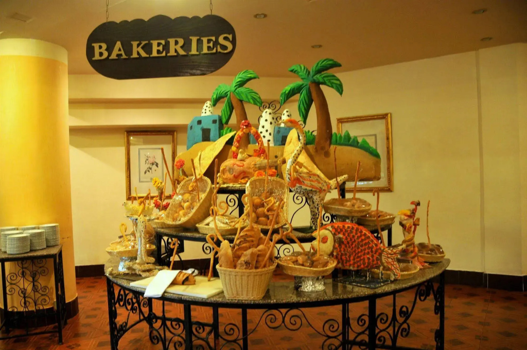 Resort Grand Oasis Resort: An indoor display area with ornate tables, colorful decorative pieces, framed artwork, and a hanging “BAKERIES” sign.