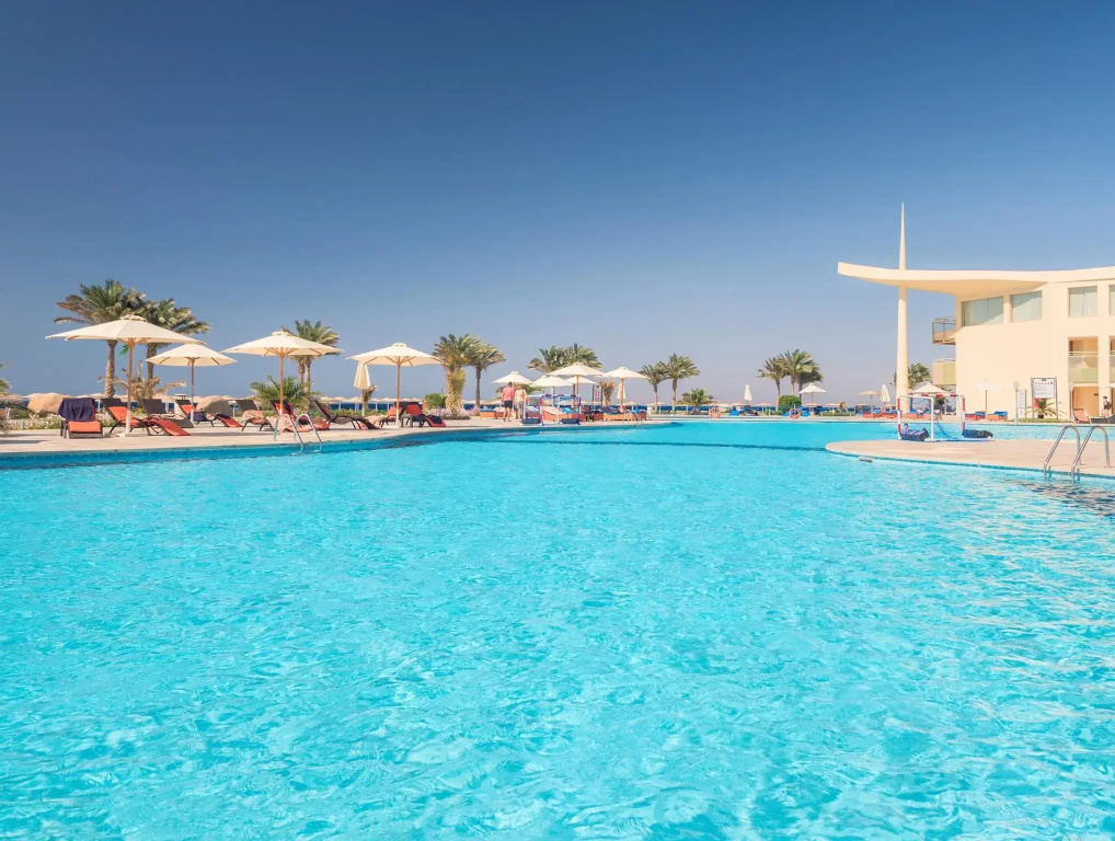 Barceló Tiran Sharm: A spacious outdoor pool is lined with sun loungers and shade umbrellas, with palm trees and the hotel building beyond.