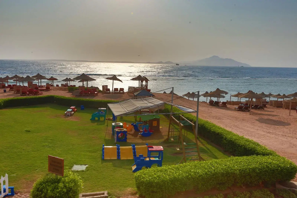 Charmillion Sea Life Resort: A beachfront lawn with a children’s playground, shaded by a canopy, overlooks the sea and rows of beach loungers and parasols.