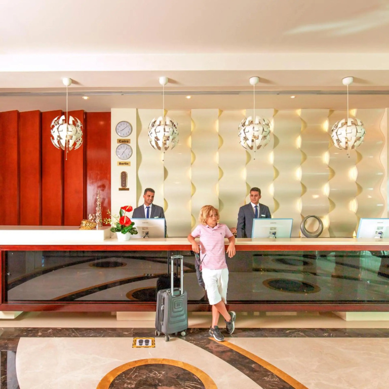 Pickalbatros Laguna Club Resort Sharm El Sheikh: A bright hotel lobby reception with a staffed front desk and a guest with rolling luggage.