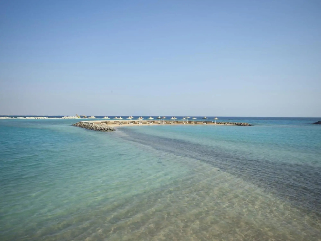 Coral Beach Resort: Calm turquoise water stretches over a sandy seabed, with a distant shoreline and open blue sky.