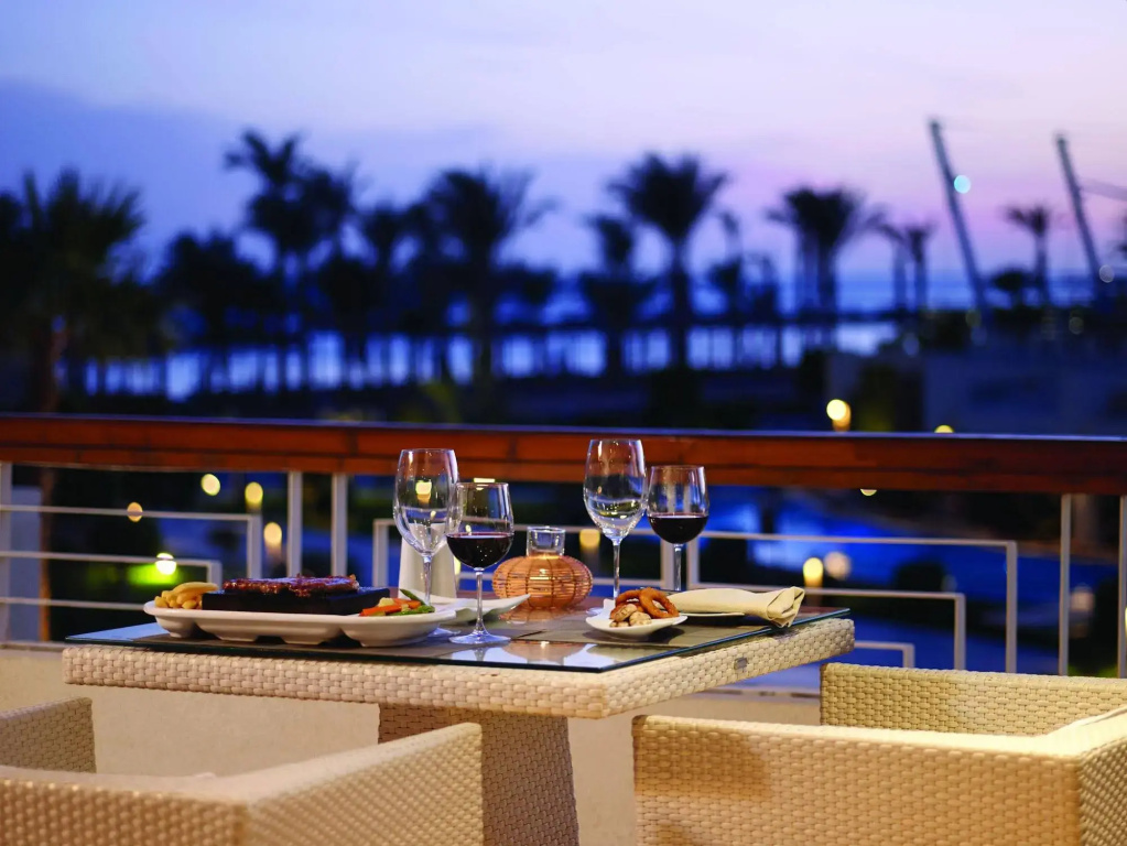 Coral Sea Sensatori Resort: An outdoor terrace table is set for dinner with plated food and wine glasses, surrounded by woven chairs and palm silhouettes at dusk.