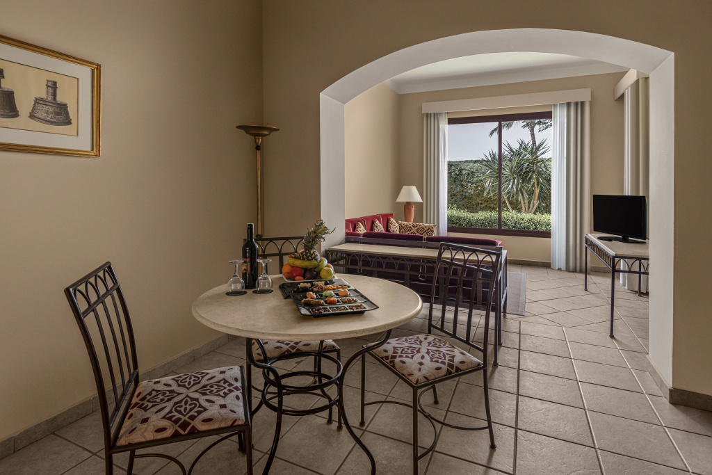JAZ Makadi Saraya Resort: A spacious living and dining area with a set dining table, sofa, television, and a large window overlooking greenery.