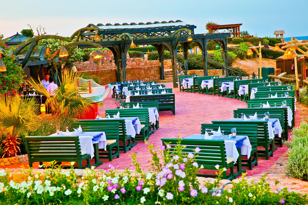 Charmillion Club Resort: An outdoor dining terrace with rows of set tables beneath a covered pergola, surrounded by flowering garden beds.