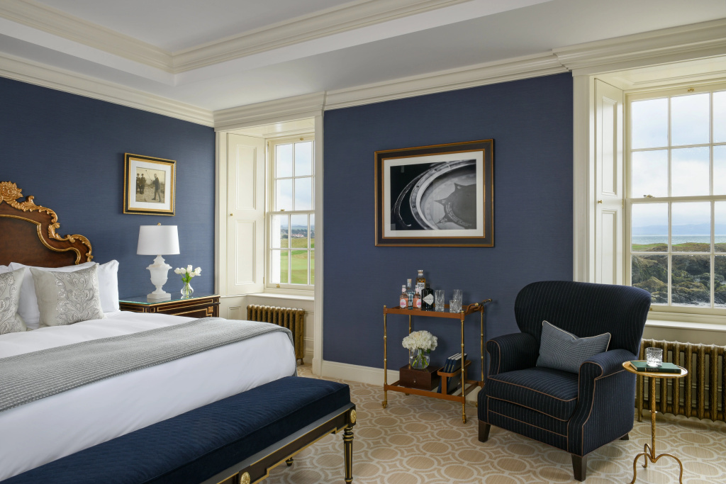 A suite bedroom with deep navy walls, a carved gilt headboard, white linen and an upholstered bench at the foot of the bed, a navy wing armchair, a small drinks table and framed pictures, with the coastline visible through the windows