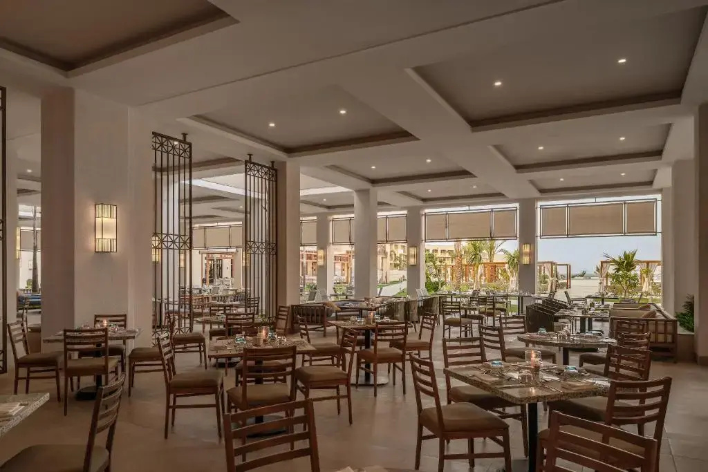 JAZ Elite Asteria Hotel: A spacious restaurant dining room with set tables, warm lighting, and large windows overlooking greenery.
