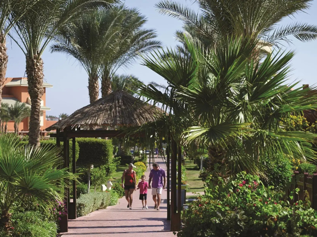 صورة: Coral sea Holiday Resort & Aqua park: A shaded garden walkway winds beneath a thatched pergola, surrounded by palms and flowering greenery.