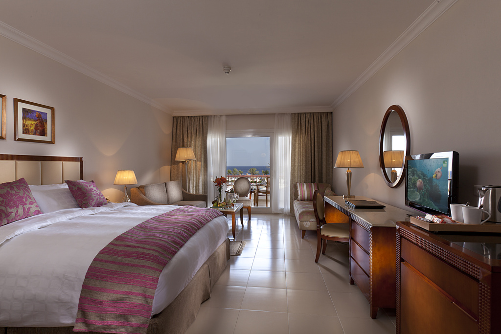 Baron Palace Sahl Hasheesh Resort: A bright bedroom with a sitting area, television, and balcony doors overlooking the sea.