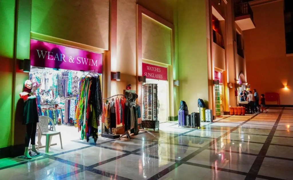 Charmillion Club Aqua Park: An indoor shopping arcade with clothing, swimwear, watch and sunglasses shops along a spacious tiled walkway.