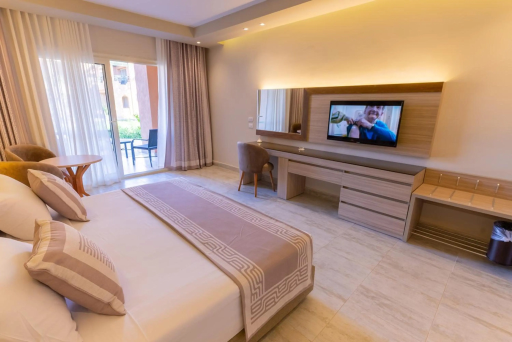 Ali Baba Palace Hotel: A bright guest room with a large bed, wall-mounted TV, desk and chair, and a balcony visible through the glass doors.