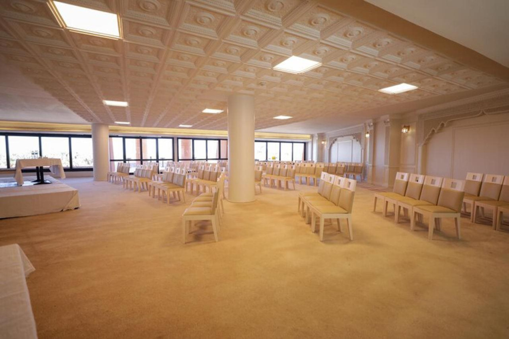 Calimera Blend Paradise: A spacious event hall is arranged with rows of chairs facing a front table, with large windows and decorative ceiling details.