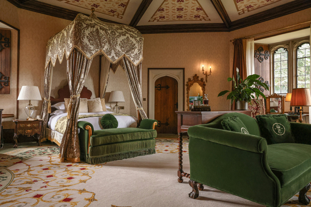 A four-poster canopy bed with richly patterned fabric drapes, a green sofa at its foot, a painted ceiling and a tall window letting in daylight