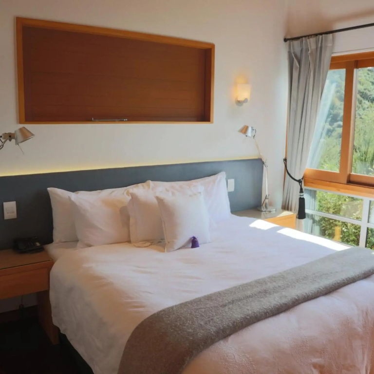 A bedroom with a white bed against a grey upholstered headboard and a timber panel above, beside a window looking onto a bush-covered hillside