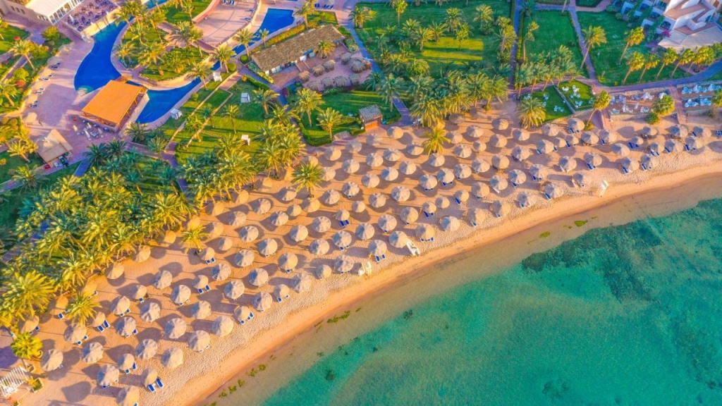 Fort Arabesque Resort, Spa & Villas: A palm-lined sandy beach is dotted with rows of thatched parasols and loungers beside clear turquoise water, with a swimming pool visible among the resort gardens.