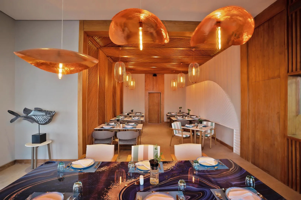 Rixos Premium Magawish Suites and Villas: An elegant indoor restaurant with set dining tables, warm pendant lighting, and natural wood details.