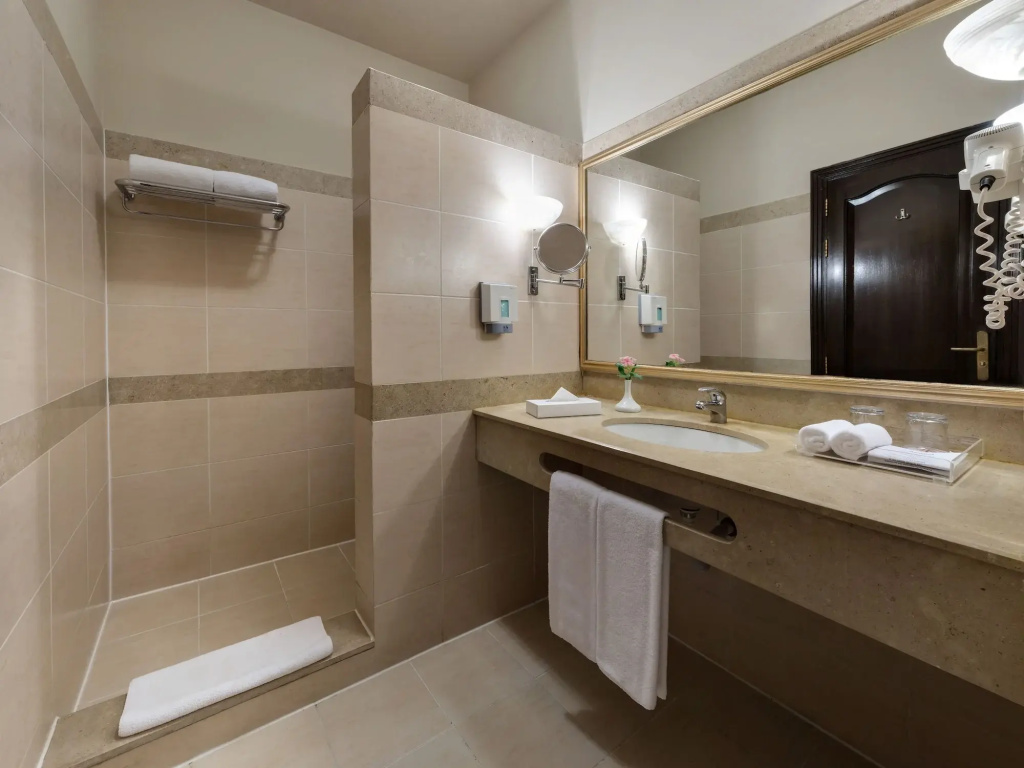JAZ Makadi Saraya Resort: A tiled bathroom with a wide mirrored vanity, sink, fresh towels, and a wall-mounted hair dryer.