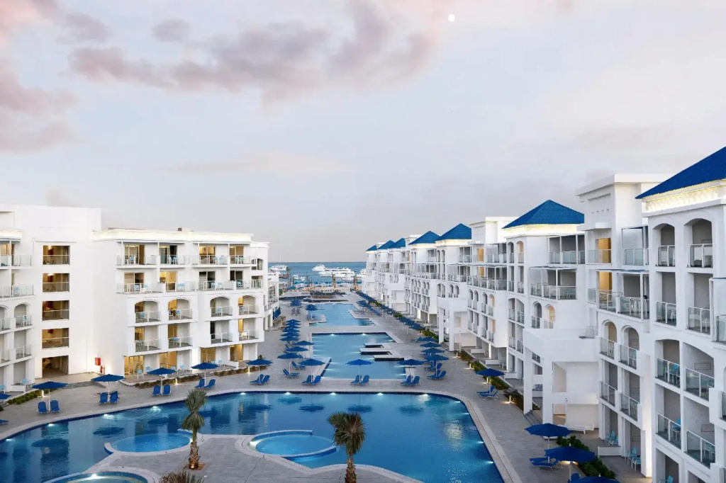 Pickalbatros Blu Spa Resort - Adults Friendly 16 Years Plus: White resort buildings with blue roofs overlook long outdoor pools lined with sun loungers and umbrellas, with the sea beyond.