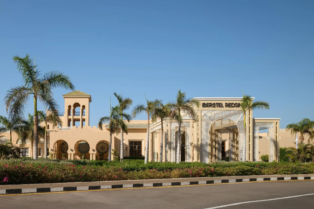TUI Magic Life Redsina: The hotel’s cream-colored facade features a prominent arched entrance, with palm trees and flower beds along the front.