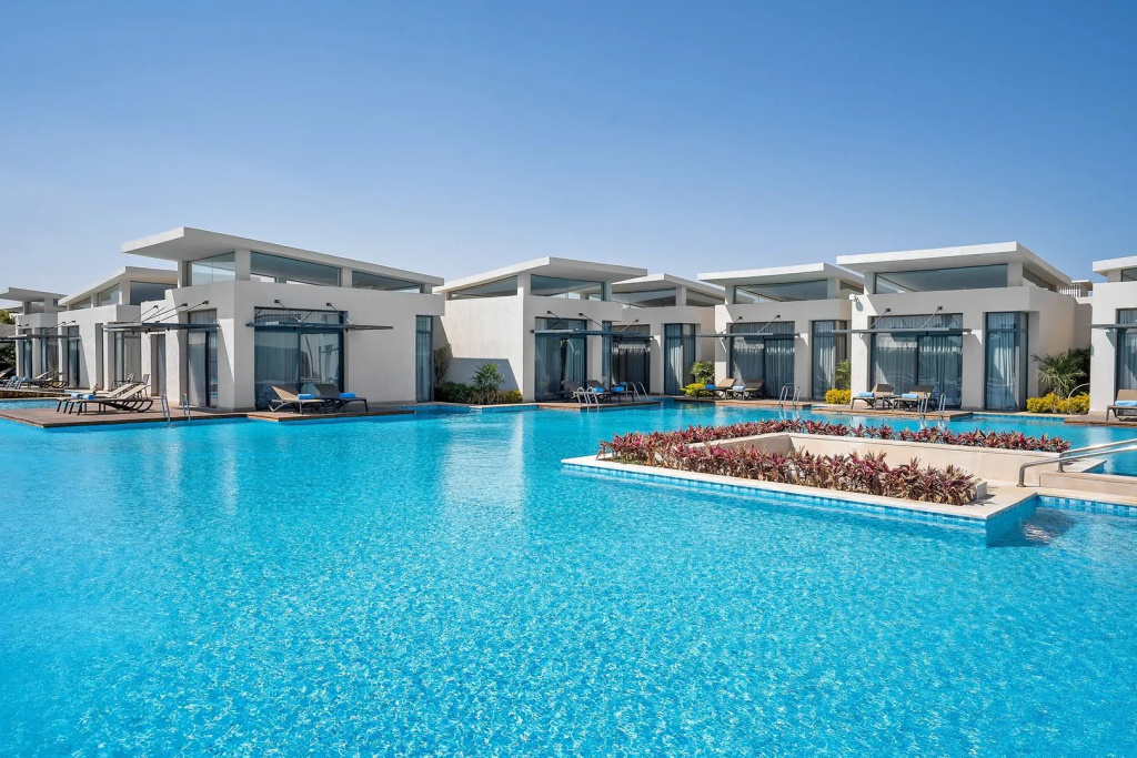 Rixos Premium Magawish Suites and Villas: Modern white suites line a large turquoise pool, with loungers beside the water and a planted island in the foreground.