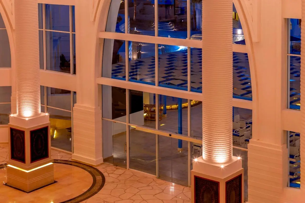 Pickalbatros Palace Resort - Sharm El Sheikh: A spacious, warmly lit interior with tall columns, expansive glass panels, and patterned tile flooring.