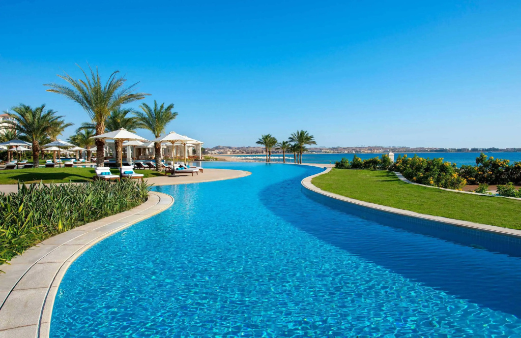 Baron Palace Sahl Hasheesh Resort: A sweeping outdoor pool is surrounded by palm trees, loungers, and shaded umbrellas, with a sea view beyond.
