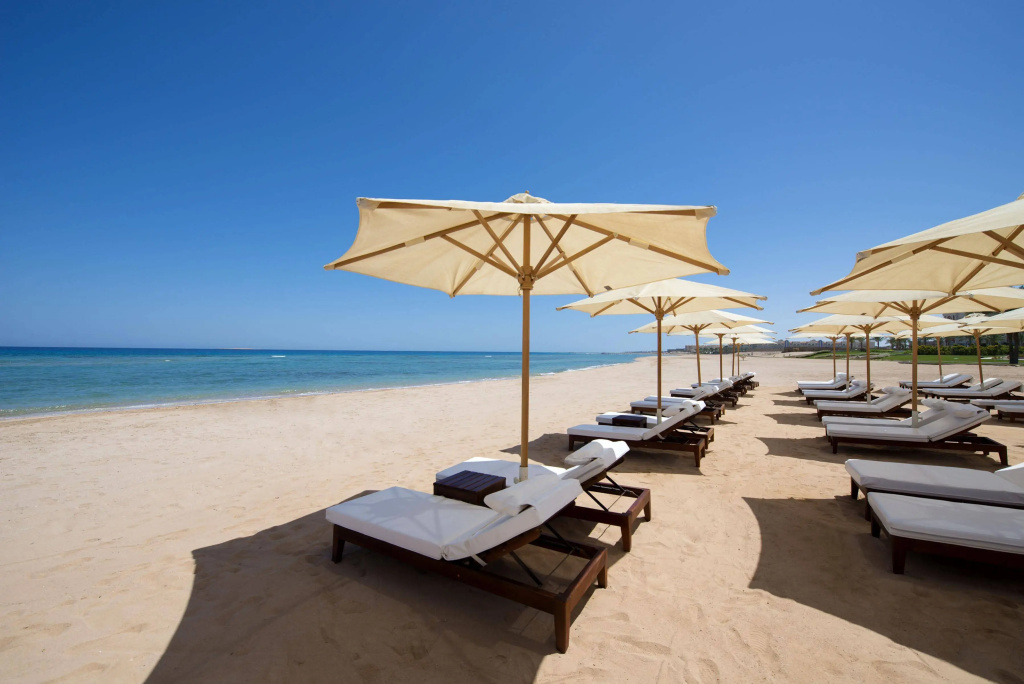 Baron Palace Sahl Hasheesh Resort: Cushioned sun loungers and large umbrellas line a sandy beach beside the calm blue sea.