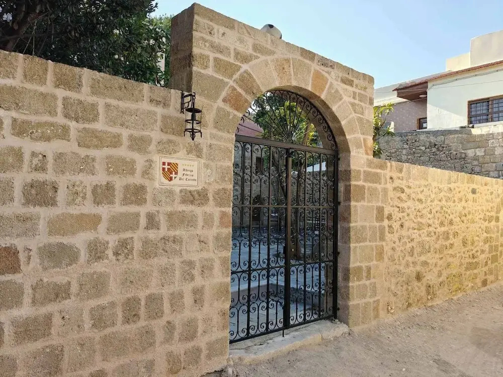 חומת אבן חול בעיר העתיקה של רודוס ובה שער קשתי עם סורג ברזל מעוטר, שלט קטן ועליו הכיתוב Albergo Fabrizio Del Carretto, ומעבר לשער נראית חצר מרוצפת