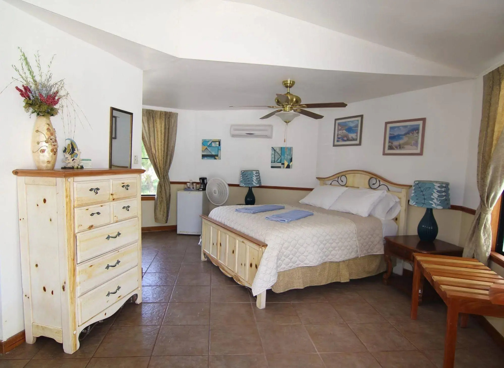 A simple guest room with white walls and a tiled floor, a pale wooden dresser, a bed with a cream quilt and folded blue towels, and a ceiling fan