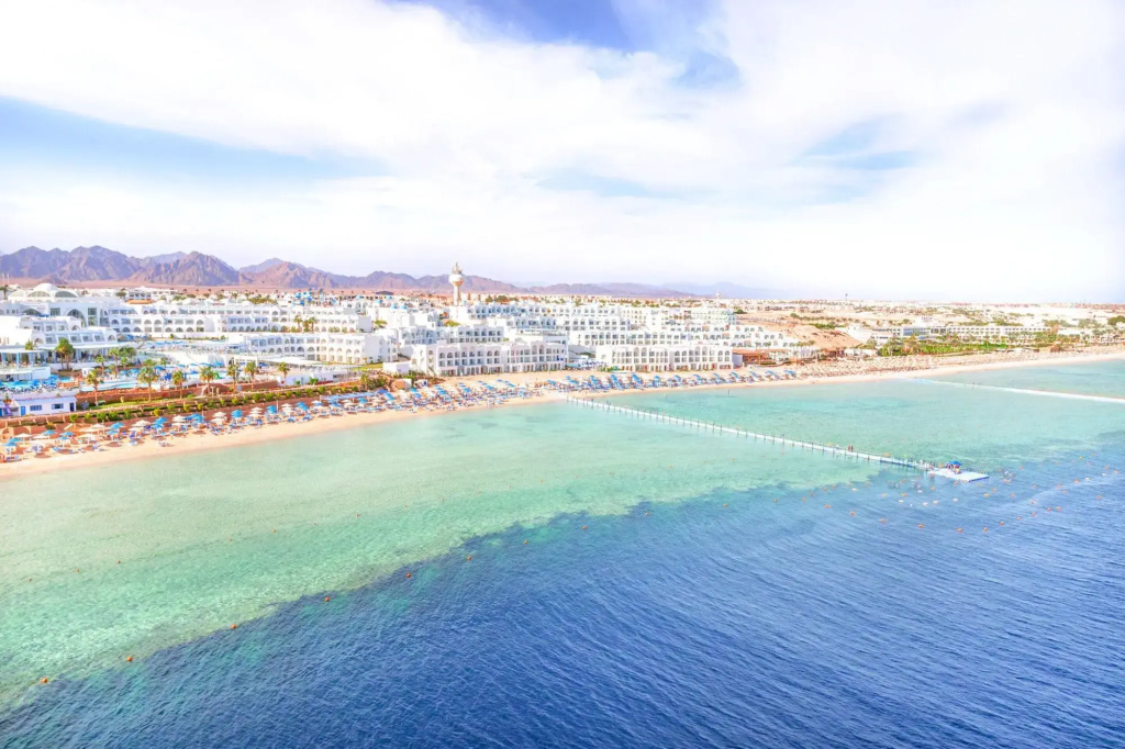 Pickalbatros Palace Sharm & Aqua Park: A beachfront resort with a sandy shore, rows of sun loungers and umbrellas, and clear turquoise water.