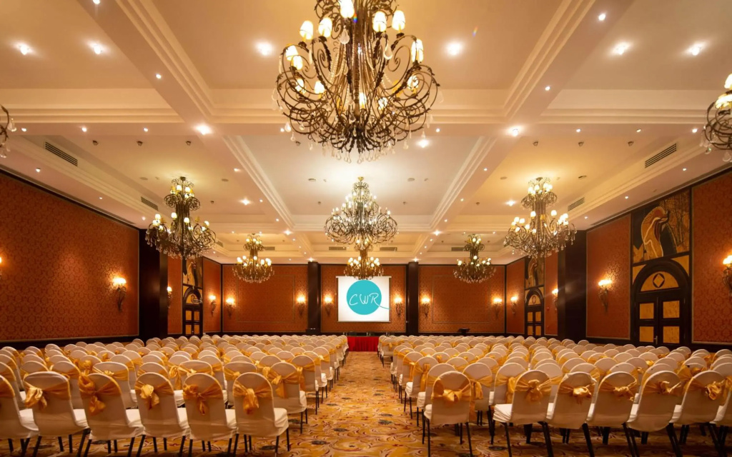 Sentido Caribbean world soma Bay: A spacious event hall with rows of chairs facing a presentation screen, beneath ornate chandeliers.