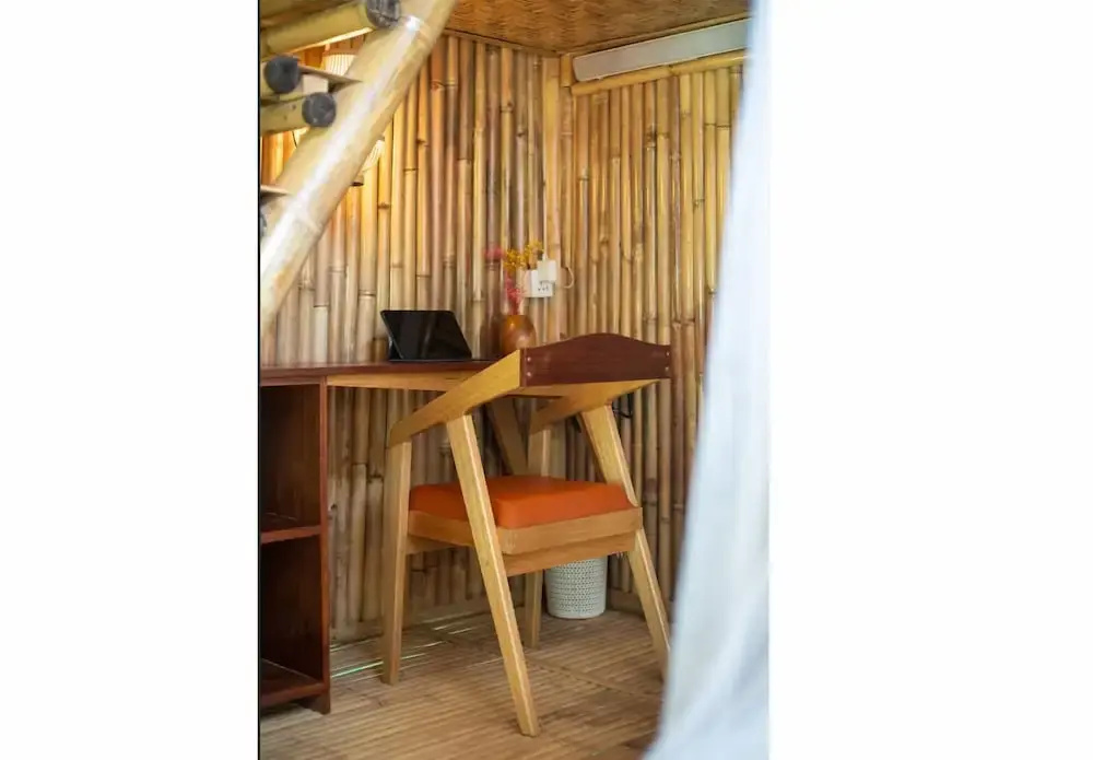 The corner of a bamboo-walled room with a wooden desk, a tablet propped open, a pale wood chair with an orange cushion and a small vase of yellow flowers