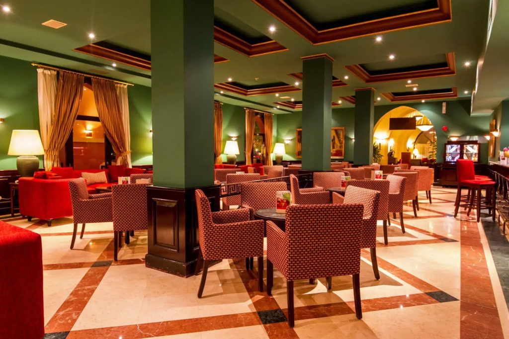 JAZ Makadi Saraya Resort: A warmly lit indoor lounge with woven armchairs, red sofas, and scattered tables.