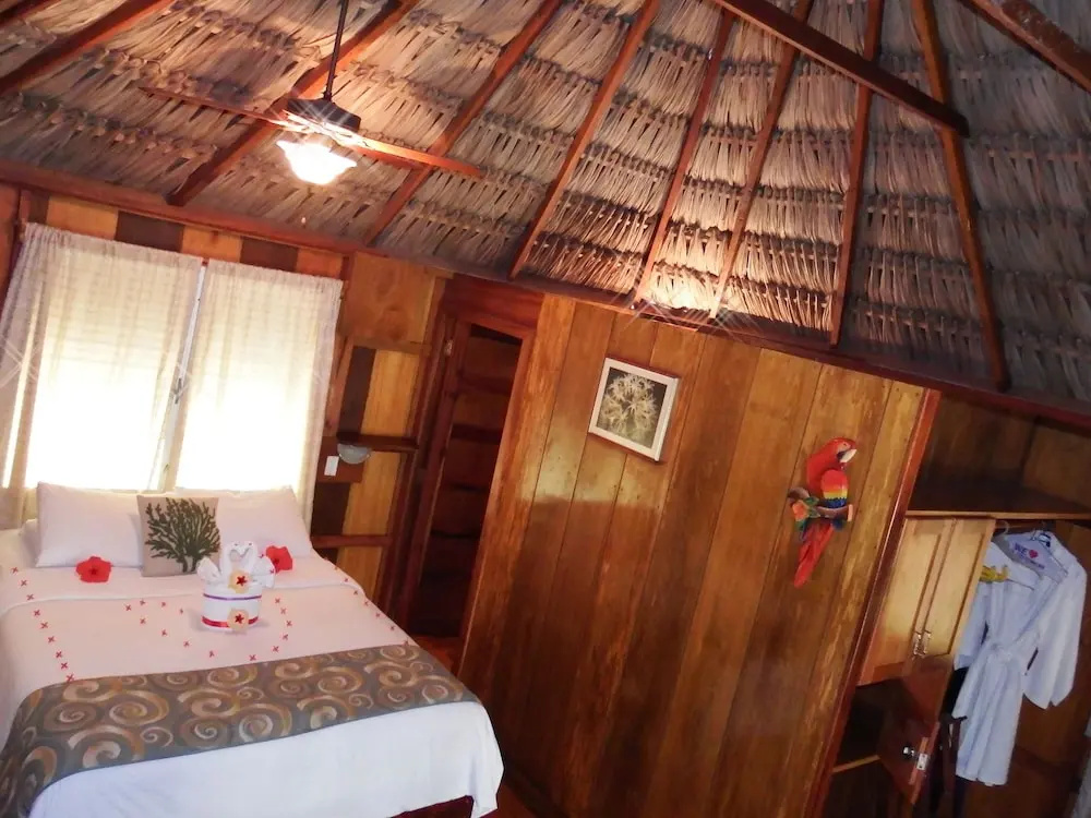 A cabana interior beneath a thatched roof with polished timber walls, a bed laid with towel swans and red flowers, and a carved parrot on the wall