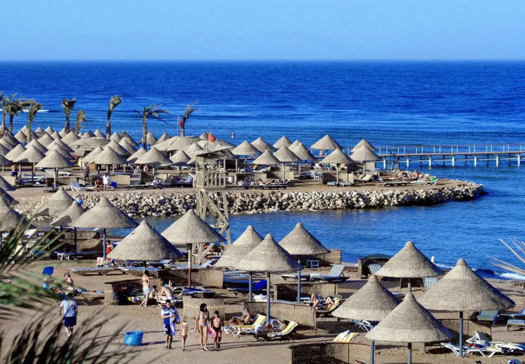 Parrotel Beach Resort: Relax on a sunny seaside beach beneath thatched parasols, with loungers and a pier extending over the blue water.