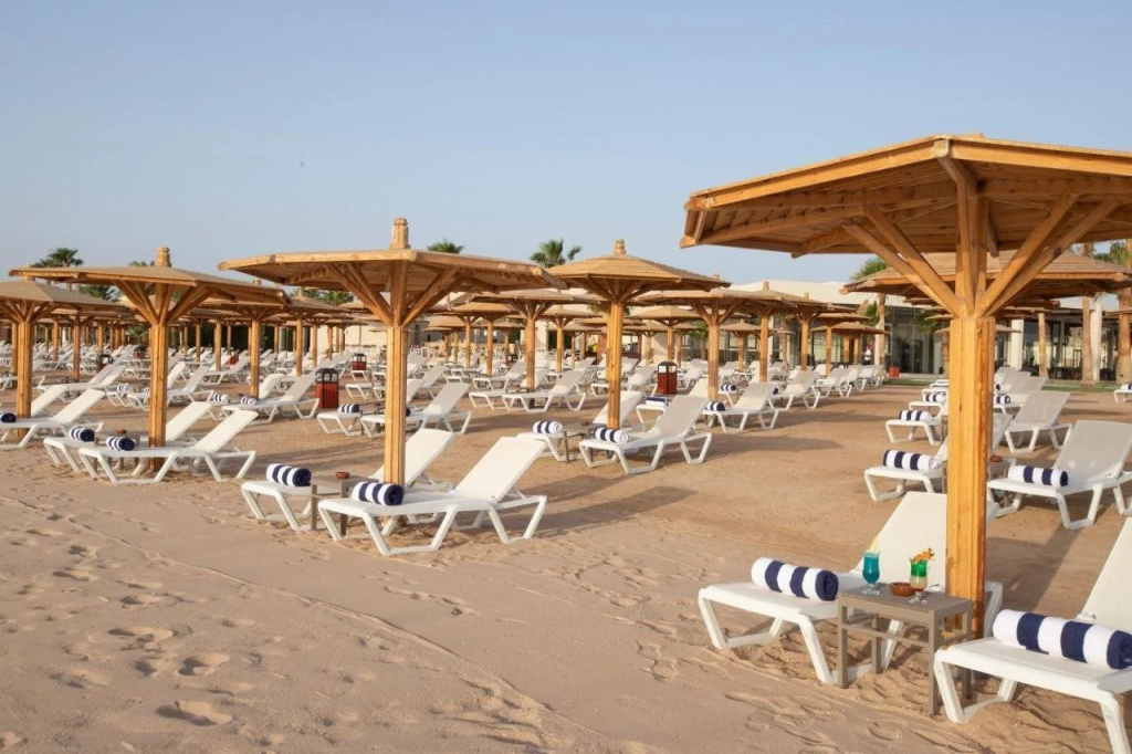 Xanadu Makadi Bay - High Class All Inclusive: White sun loungers with striped towels sit beneath wooden shade canopies on a sandy beach.