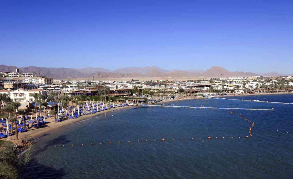 Lido Sharm Hotel Naama Bay: A sunny beachfront scene with rows of loungers along a curved shore, overlooking a calm blue bay and distant mountains.