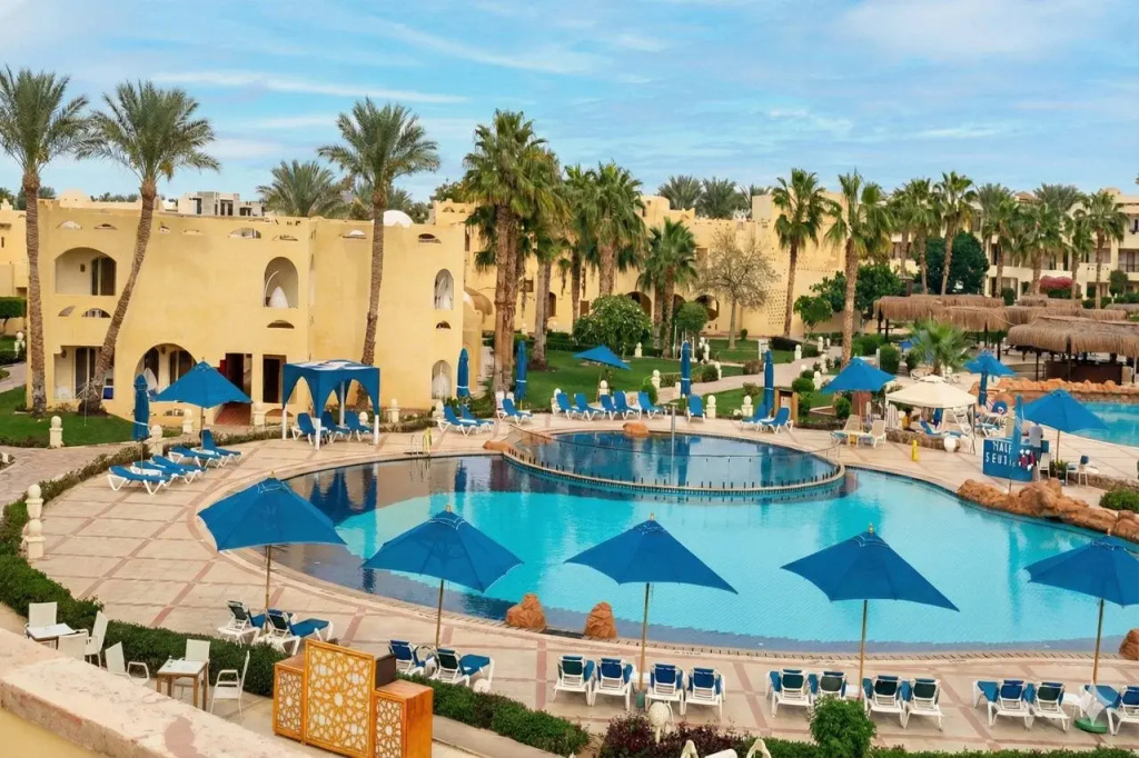 Regency Plaza Aqua Park & SPA: A large outdoor pool is surrounded by sun loungers and blue parasols, with palm trees and resort buildings beyond.