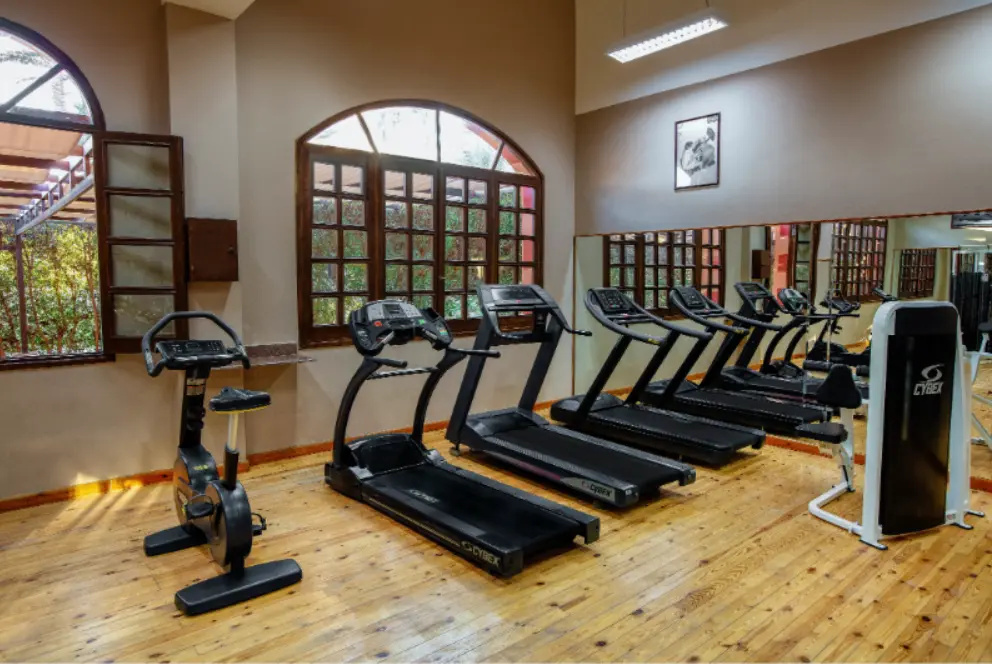 JAZ Makadi Oasis Resort: A bright fitness room with several treadmills and cardio machines, wood-look flooring, and tall arched windows.