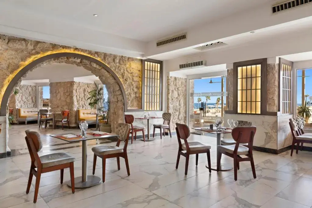 Pickalbatros Citadel Resort: A bright dining area with tables and cushioned chairs, framed by large windows and a stone arch.