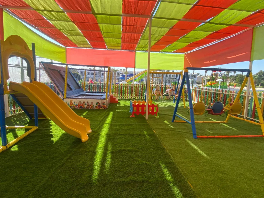 Invidia Coral Beach Tiran: A shaded outdoor playground with slides, swings, and artificial turf.
